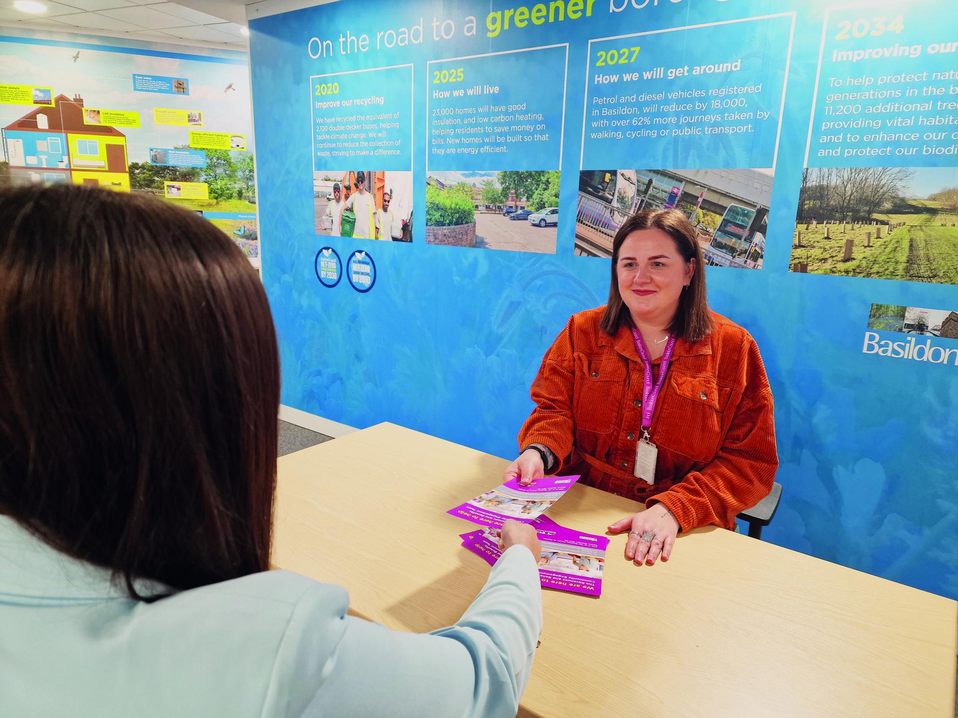 A woman sitting at a desk handing a leaflet to someone